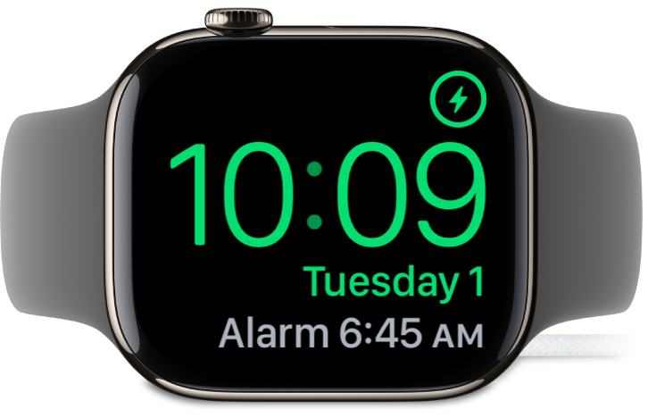 An Apple Watch placed on its side and connected to the charger, with the screen showing the charging symbol in the top-right corner, the current time below that, and the time of the next alarm.