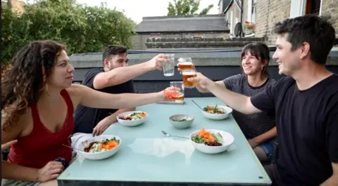 BBC Two couples clink glasses and sit at a table eating food in the garden. The two men wear dark t-shirts. One woman wears a maroon top and the other wears a stripy T-shirt.