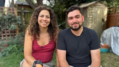 A couple in their thirties sit next to each other in the garden, smiling at the camera. The brunette man wear a dark t-shirt and the woman wears a maroon vest top.