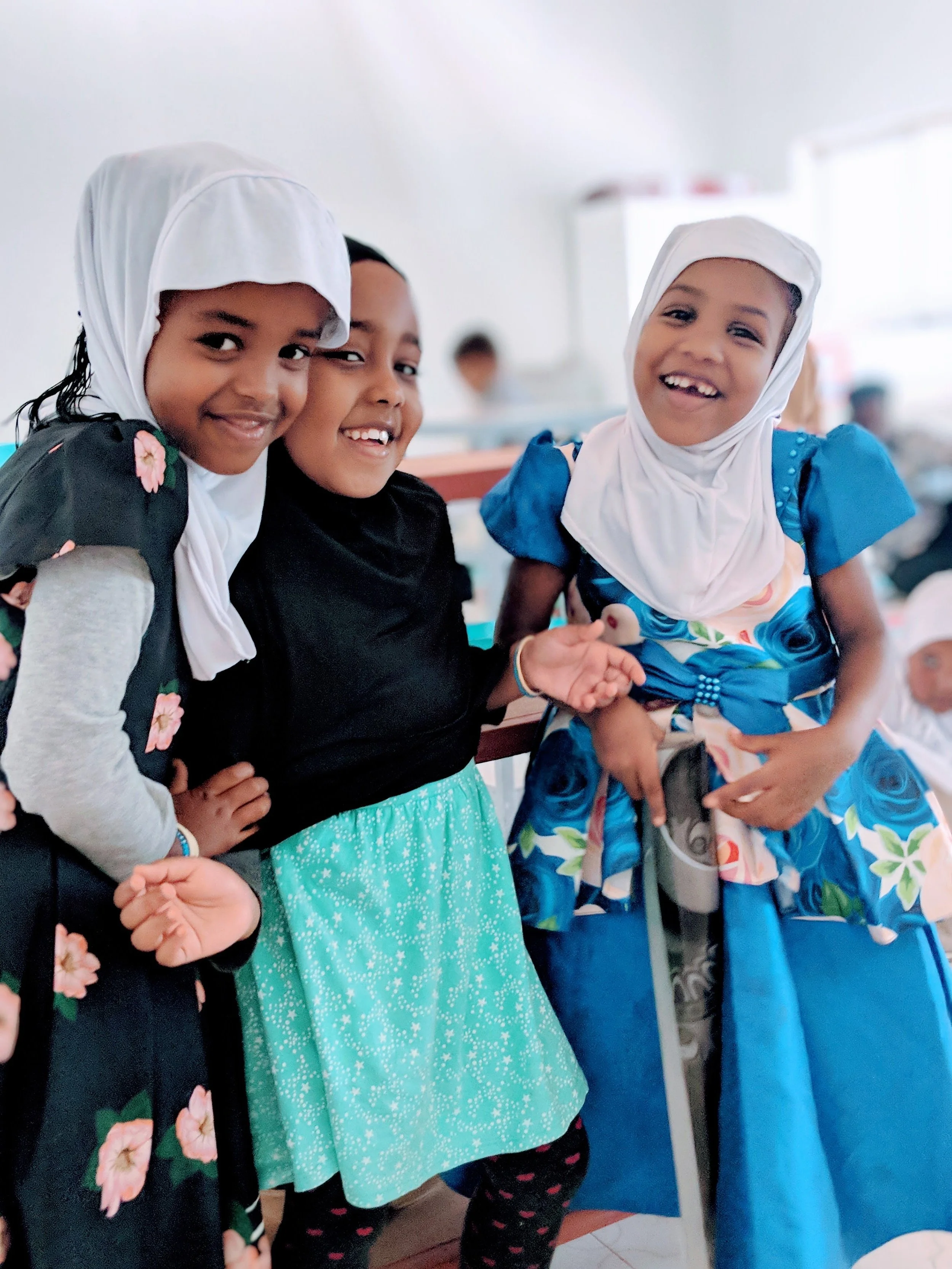 Three young girls wearing hijabs and colorful dresses smiling and posing together in a classroom setting.