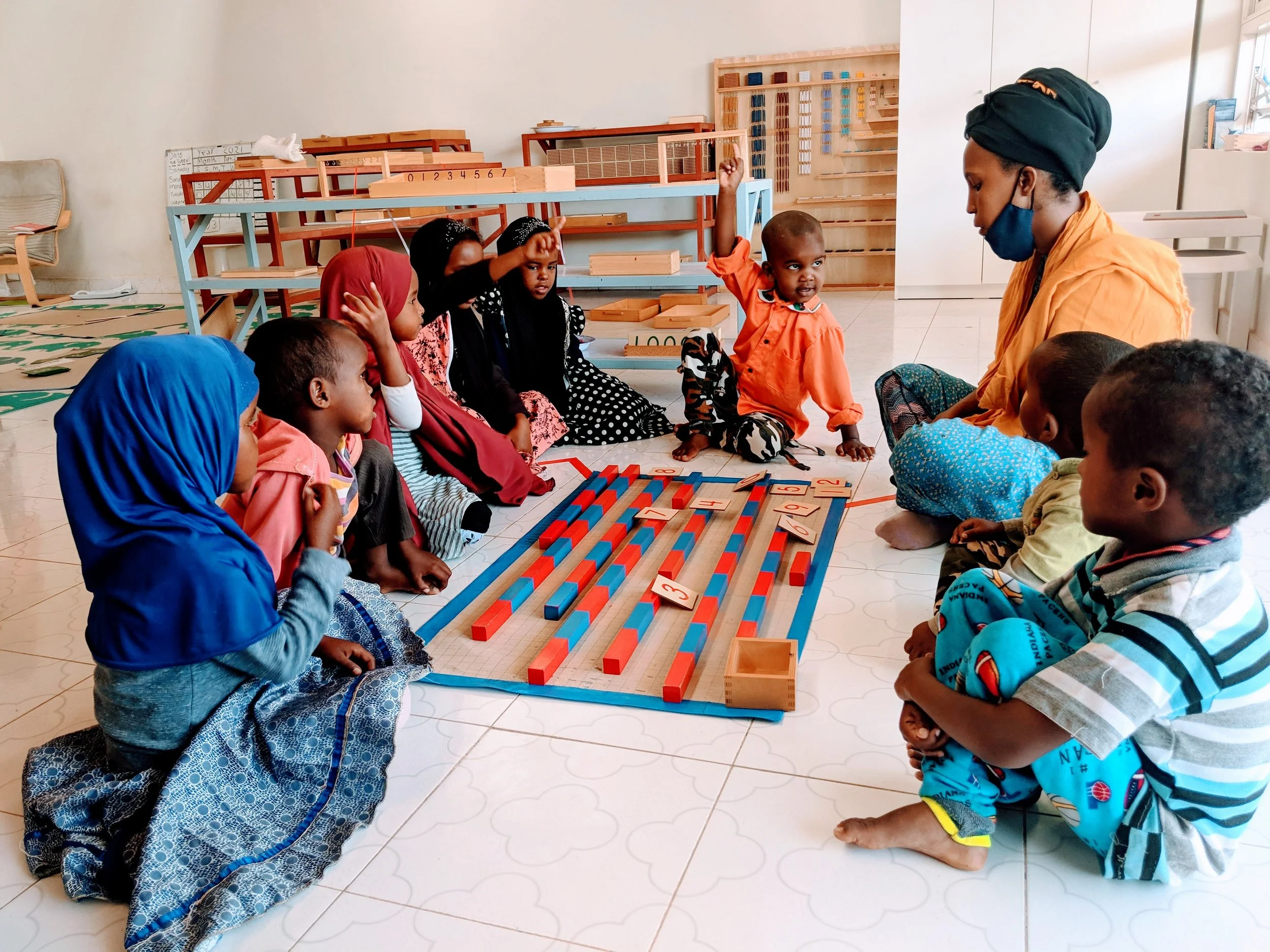Young children sitting on the floor in a classroom, engaged in an educational activity with an instructor. There are colorful number blocks and a grid on the floor, with educational materials in the background.