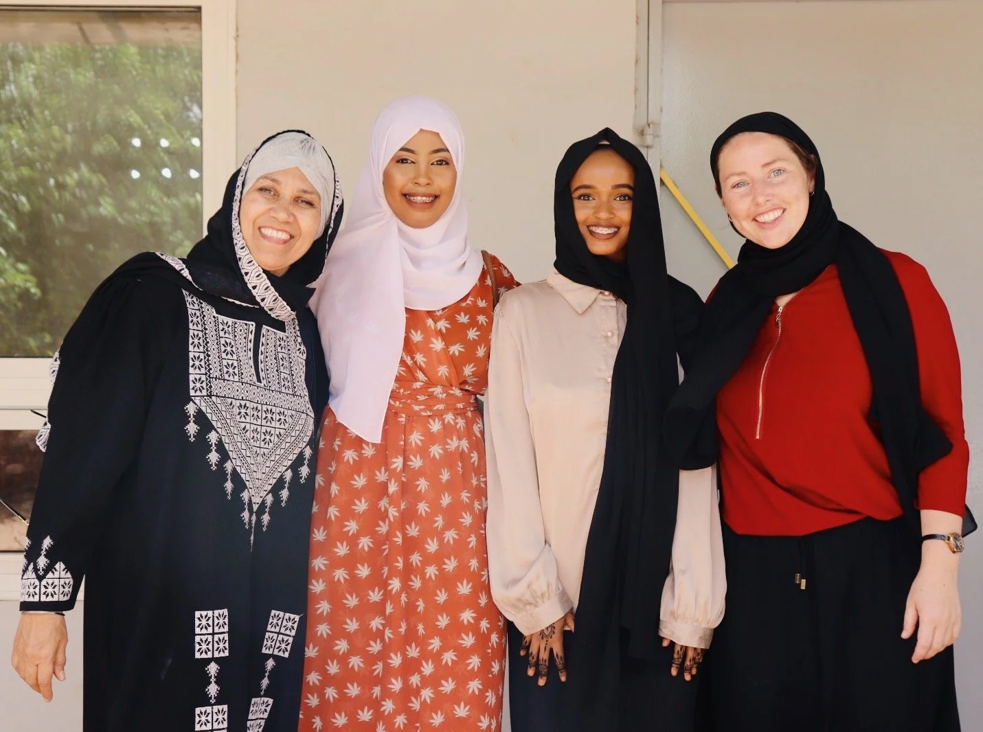 Four women standing side by side, smiling, dressed in modest clothing and wearing headscarves in different styles and colors, with a door and window in background.