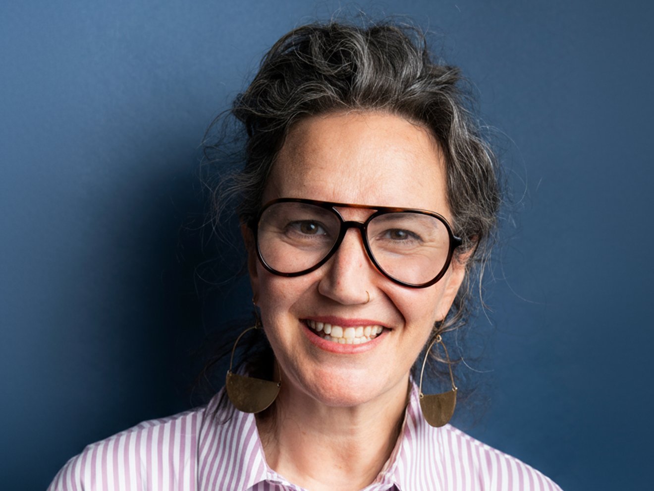 A headshot of a woman with gray-flecked hair, large dark-rimmed glasses, and gold crescent earrings, smiling against a solid blue background.