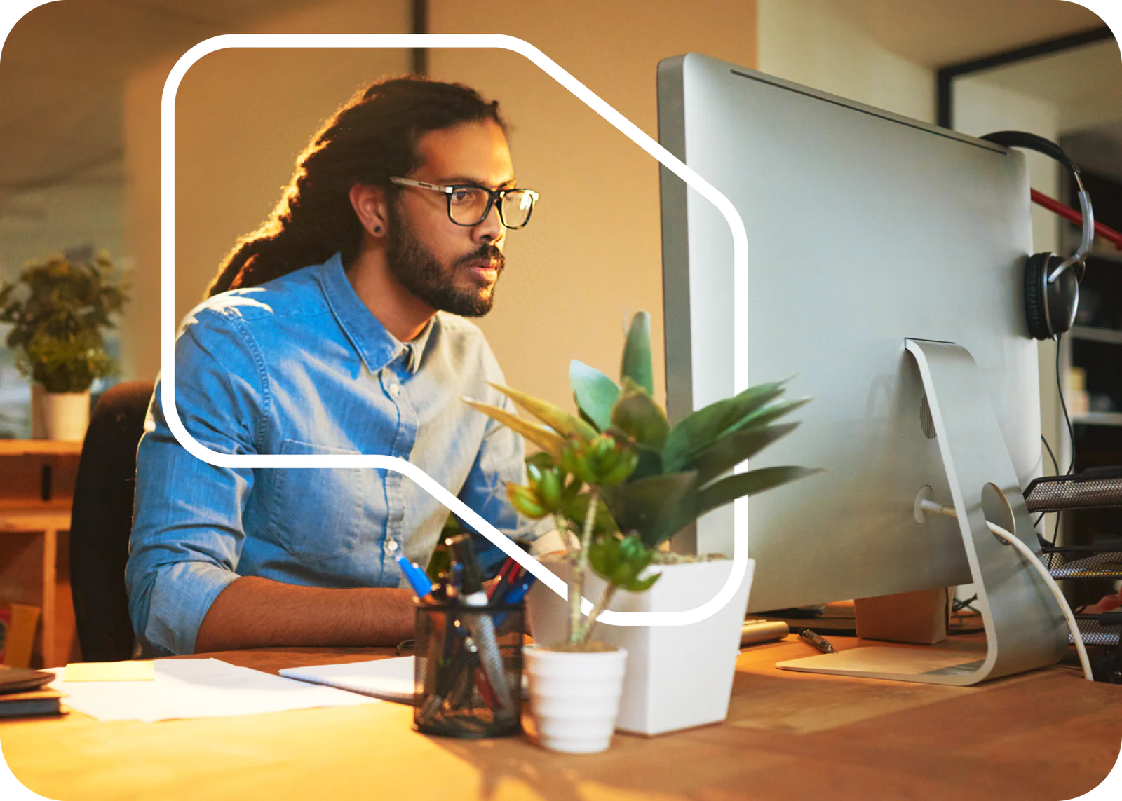 Man with glasses and dreadlocks working at a desktop computer in an office with plants.